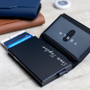 May include: A black wallet with the name "Sean Taylor" in white script is open, revealing credit cards. A clear card holder with the Tesla logo is visible. Two other wallets, one blue and one brown, are stacked in the background.
