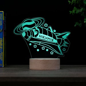 May include: A teal blue night light shaped like a rocket ship flying through space with a planet and stars. The rocket ship has the name "BENJAMIN" written on it. The night light is on a wooden base.