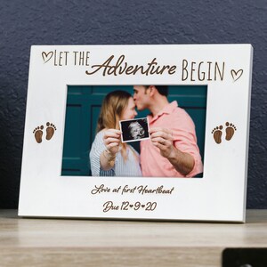 May include: White wooden picture frame with the words "LET THE Adventure BEGIN" and "Love at first Heartbeat" engraved. The frame holds a sonogram image and has baby footprints. The date "Due 12-9-20" is also engraved.