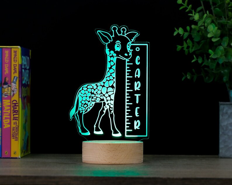 May include: A turquoise blue giraffe night light with a wooden base. The giraffe is holding a ruler with the name "Carter" on it.