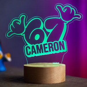 May include: A neon green LED night light with the name "Cameron" and a "hang loose" hand gesture design. The light sits on a round wooden base. The background is a gradient of purple and black.