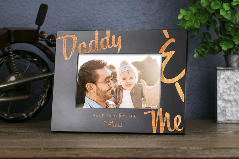 以下が含まれることがあります： 「Daddy & Me Best Pals for Life Olivia」と金色の文字で書かれた黒い写真フレーム。フレームには、父親と子供の2人の写真が入っています。