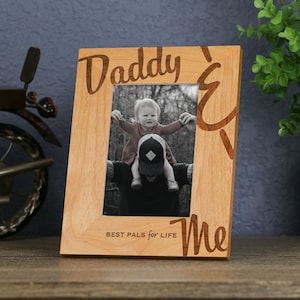 以下が含まれることがあります： 前面に「Daddy & Me Best Pals for Life」という文字が刻まれた木製の写真フレーム。フレームには、白黒の父親と子供の画像が入っています。