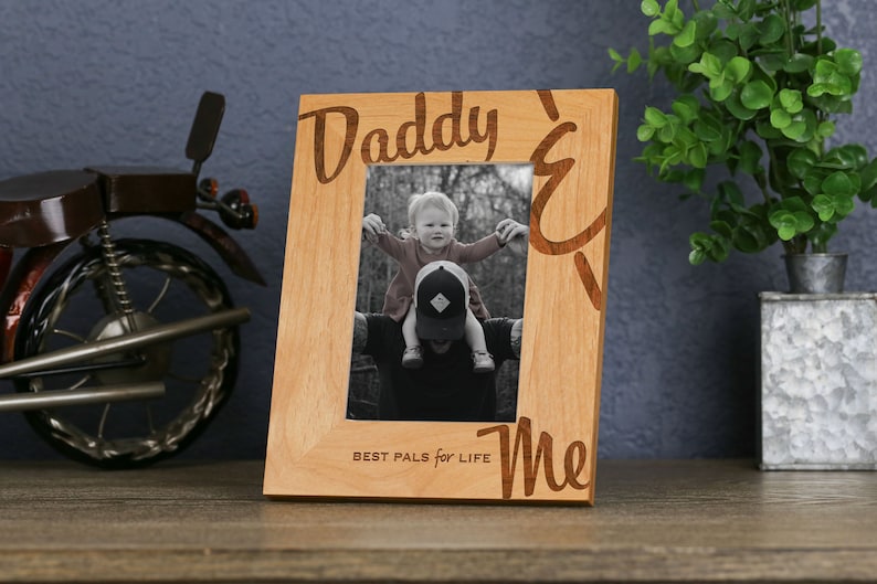 以下が含まれることがあります： 前面に「Daddy & Me Best Pals for Life」という文字が刻まれた木製の写真フレーム。フレームには、白黒の父親と子供の画像が入っています。
