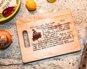 May include: A wooden cutting board with a handwritten red cake recipe. The recipe includes ingredients like sugar, shortening, eggs, flour, cocoa, salt, vanilla, and baking soda. The board has a handle and is engraved with a small image of a cart.