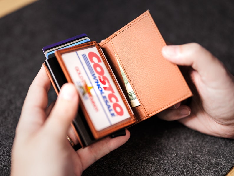 May include: A brown leather wallet with a Costco Wholesale membership card inside. The wallet is open and there is a bill of cash inside.