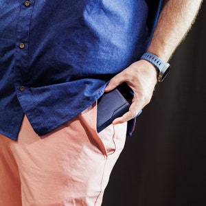 May include: A person wearing a blue button-down shirt and pink shorts is holding a black wallet in their right hand.