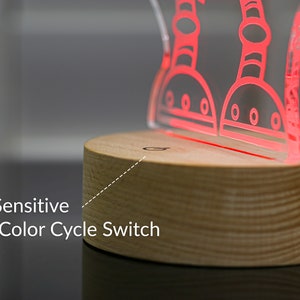 May include: A wooden base with a touch-sensitive power and color cycle switch. Atop the base is a clear acrylic lamp with a red outline of two robots.