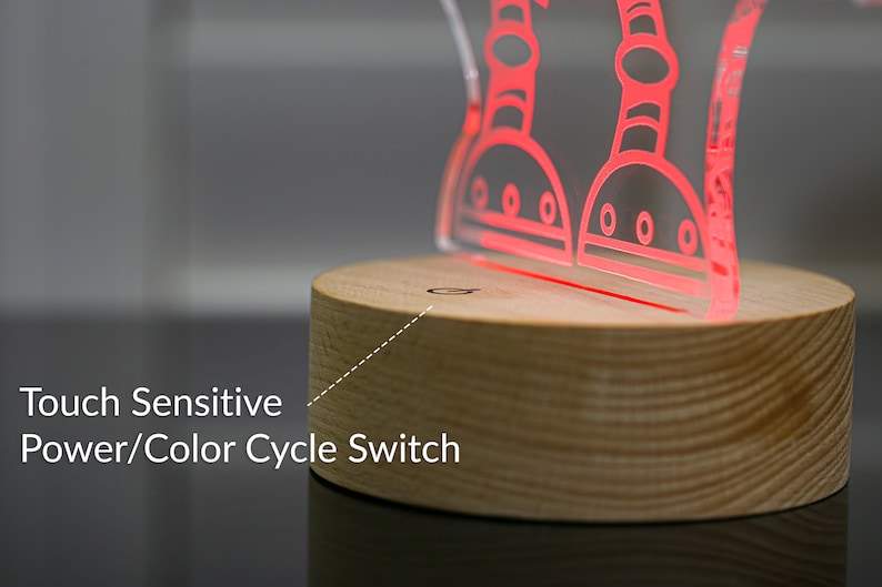 May include: A wooden base with a touch-sensitive power and color cycle switch. Atop the base is a clear acrylic lamp with a red outline of two robots.