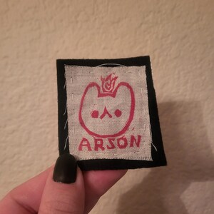 May include: A black and white patch with a red cartoon animal with a flame on its head. The text "ARSON" is printed below the animal.