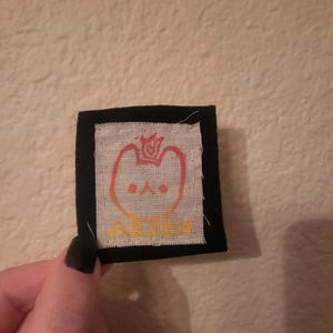 May include: A black fabric patch with a white fabric square in the center. The white fabric square has a red, orange, and yellow gradient design of a cartoon cat with a crown on its head. The text "ARION" is written in yellow below the cat.