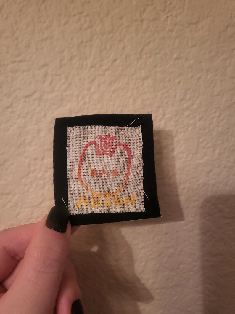 May include: A black fabric patch with a white fabric square in the center. The white fabric square has a red, orange, and yellow gradient design of a cartoon cat with a crown on its head. The text "ARION" is written in yellow below the cat.