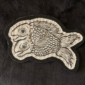 May include: A black fabric patch with a white embroidered design of two fish with scales. The fish are facing each other with their mouths open.