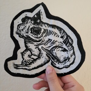 May include: Black and white linocut print of a frog on a white fabric patch with a black border. The frog is sitting on its hind legs with its front legs tucked in.
