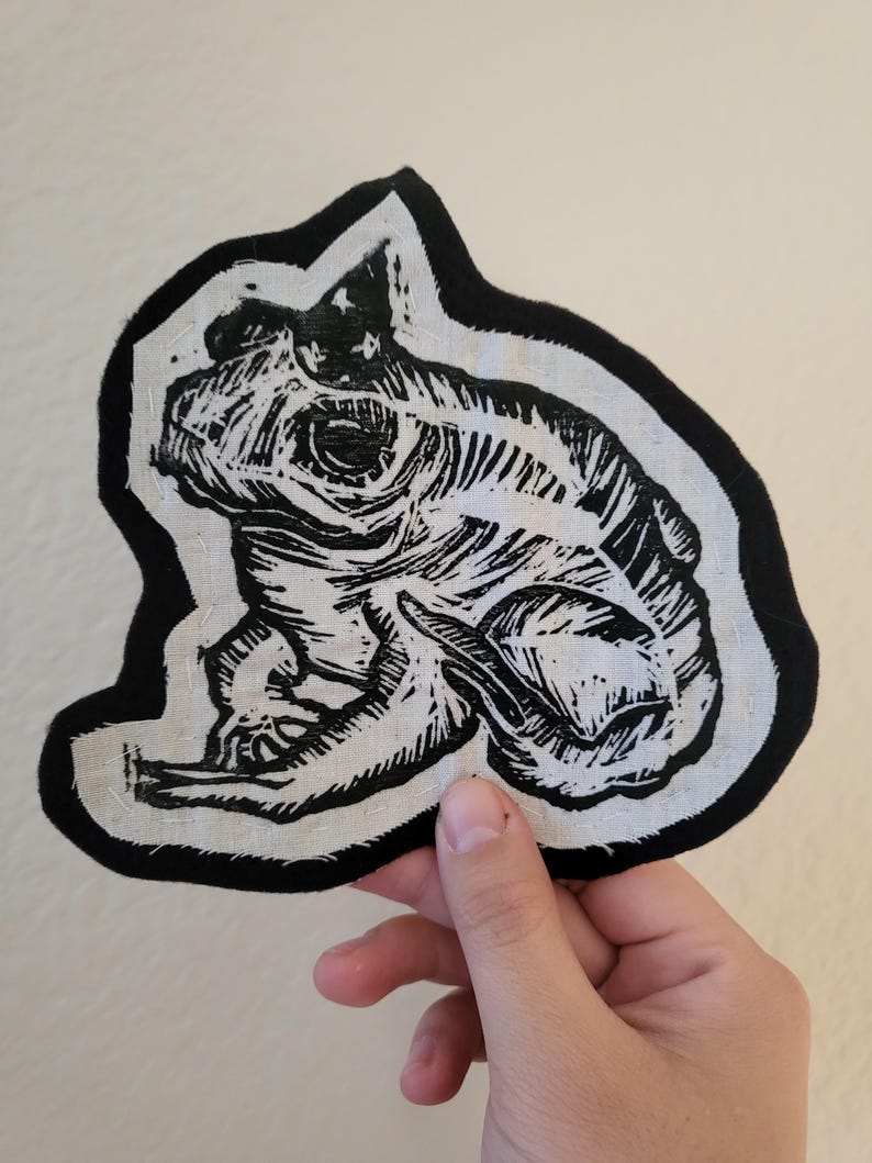 May include: Black and white linocut print of a frog on a white fabric patch with a black border. The frog is sitting on its hind legs with its front legs tucked in.