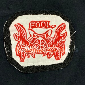 May include: A red and white fabric patch with a stylized crab design and the word "FOOL" printed above it. The patch is surrounded by a black fabric border.