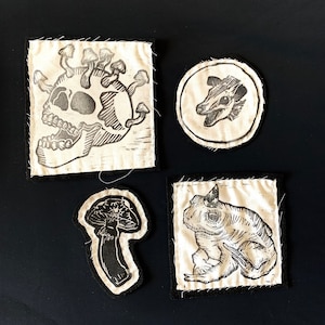 May include: Four embroidered patches with black and white designs on a white fabric background. The designs include a skull with mushrooms growing out of it, a frog, a mushroom, and a possum head.