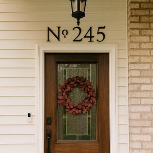 May include: A wooden front door with a glass panel and a burgundy wreath. Above the door are the black metal house numbers "No. 245". A black lantern hangs from the ceiling. The house exterior is painted cream with white trim and a brick accent.