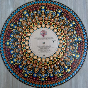 Può includere: Un disco in vinile circolare decorato con un motivo mandala dipinto a mano a cerchi concentrici con punti nei toni del blu, oro, rosso e marrone chiaro. L'etichetta centrale mostra testo tra cui Emmylou Harris, Evangeline e la lista tracce.