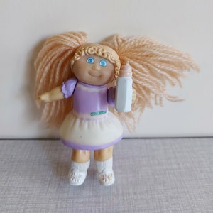 Peut inclure: Poupée Cabbage Patch Kids vintage avec des cheveux blonds en fil de laine en couettes, des yeux bleus et une robe violet clair. La poupée tient un biberon et porte des chaussettes et des chaussures blanches. La poupée a une expression souriante.
