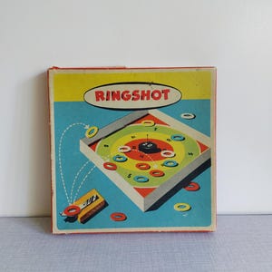 Könnte beinhalten: Vintage "Ring Shot" Spielbox mit farbenfrohem Ziel-Design. Die Box zeigt den Namen des Spiels in einem roten Oval, einen blauen Hintergrund und ein Holz-Ringwurfspiel mit bunten Ringen. Ein klassisches Spielzeug.