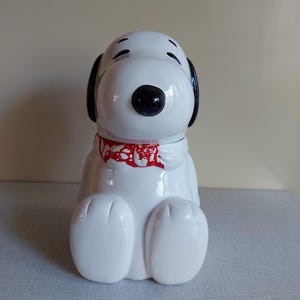 Op de afbeelding: Een witte Snoopy koekjespot van keramiek met zwarte oren, ogen en neus, en een rode kraag. De pot zit in een zittende positie met uitgestrekte benen. De pot heeft een glanzende afwerking.