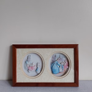 Może przedstawiać: Oprawione dzieło sztuki z dwiema okrągłymi ilustracjami królików w scenie z książki z bajkami. Rama jest w kolorze ciemnego drewna, a ilustracje mają miękkie pastelowe kolory. Dekoracja pokoju dziecięcego.