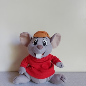 Può includere: Un topo di peluche con grandi orecchie, che indossa un maglione rosso e un berretto marrone. Il topo ha un'espressione amichevole con un ampio sorriso e denti visibili. Il giocattolo è grigio e rosso.