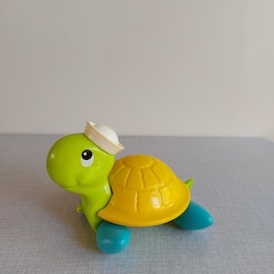 Puede incluir: Una tortuga de juguete verde y amarilla con aletas azules y un gorro de marinero blanco. La tortuga tiene un caparazón amarillo y una cara sonriente. Juguete para niños.