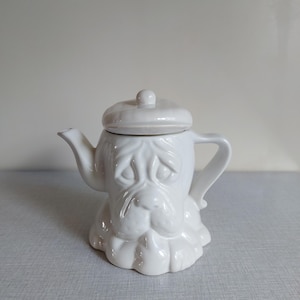 Könnte beinhalten: Eine weiße Keramik-Teekanne in Form eines Hundegesichts, mit Deckel und Henkel. Die Teekanne hat ein detailliertes, geformtes Design der Gesichtszüge eines Hundes, einschließlich Schnauze, Augen und Ohren. Ein einzigartiger und dekorativer Küchenartikel.