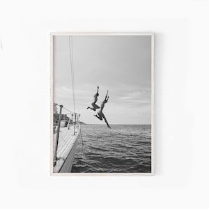 May include: Two people jump off the back of a boat into the water. The boat is white and has a wooden deck. The water is blue and the sky is cloudy.