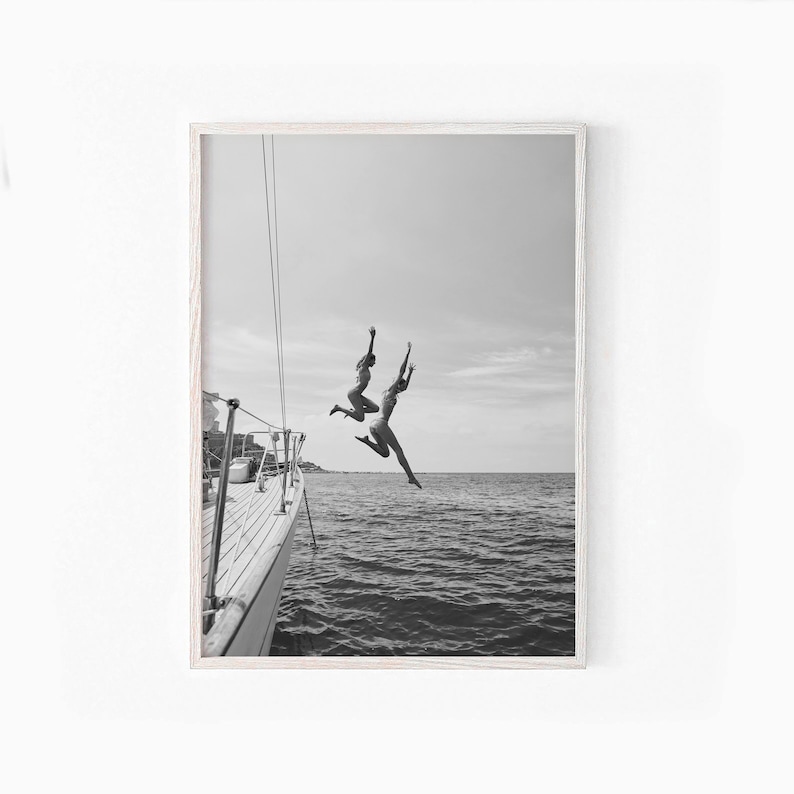 May include: Two people jump off the back of a boat into the water. The boat is white and has a wooden deck. The water is blue and the sky is cloudy.