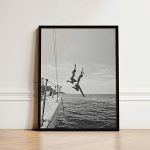 May include: Two people jump off the back of a boat into the water. The boat is white and the water is blue. The sky is cloudy and the photo is in black and white.
