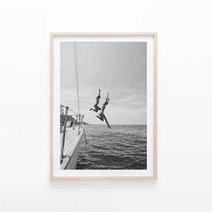 May include: Two people jump off the back of a boat into the water. The boat is docked and the water is calm. The photo is in black and white.