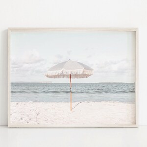 Puede incluir: Una impresi&oacute;n enmarcada de una escena de playa. Una sombrilla a rayas grises y blancas con flecos se alza sobre una playa de arena, con el mar y el cielo al fondo. La impresi&oacute;n tiene una paleta de colores suaves y apagados.