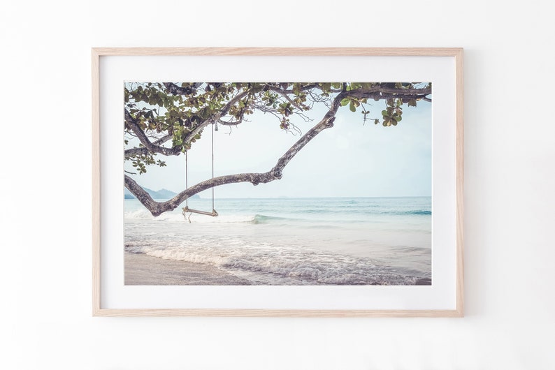 May include: A wooden swing hangs from a tree branch over a white sandy beach with turquoise water and foamy waves.