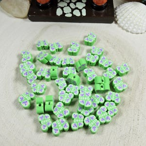 May include: A collection of green polymer clay butterfly beads with purple accents. The beads are scattered on a light wood surface, with a few cylindrical beads visible. The image also shows a decorative wooden stand with orange columns.