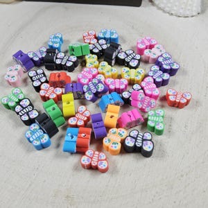 May include: A collection of colorful butterfly-shaped polymer clay beads in various colors, including pink, blue, green, yellow, and black. The beads are scattered on a light wood surface, with a seashell and decorative elements in the background.