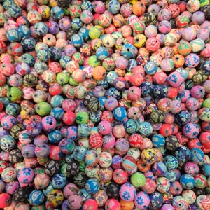 May include: A close-up shot of a large pile of colorful, patterned beads. The beads are round and feature various floral and abstract designs in shades of pink, blue, green, orange, and black. The beads are likely for jewelry making.