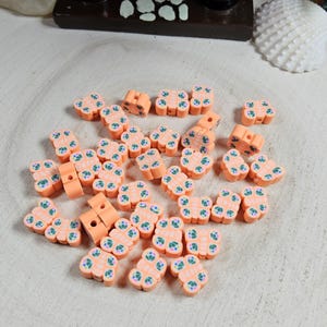 May include: A collection of small, peach-colored polymer clay beads with a floral design in blue and green. The beads are scattered on a light-colored wooden surface. Some beads have a rectangular shape with a hole.