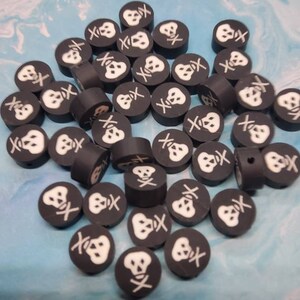 May include: A collection of black, round beads with a white skull and crossbones design. The beads are scattered on a blue and white marbled surface, suitable for jewelry making or crafting projects.