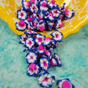 May include: A collection of small, handmade flower beads in a yellow bowl. The beads feature a white base with blue edges and pink centers. The bowl is yellow with white splatter paint.