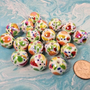 May include: A collection of round white ceramic beads featuring colorful hand-painted fruit motifs including strawberries, oranges, and watermelons, arranged next to a copper-colored United States penny for scale reference.