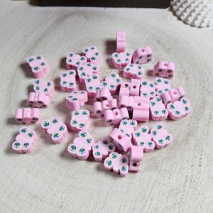 May include: A collection of pink polymer clay beads with a floral design and green accents. The beads are scattered on a light-colored wooden surface. Some beads are cylindrical, while others are flower-shaped.