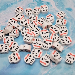 May include: A collection of white skull-shaped beads with black and red accents. Each bead features a skull design with eye sockets, a nose, and a mouth. The beads are scattered on a blue and white surface, suitable for jewelry making.