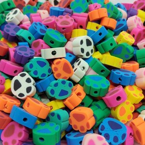 May include: A collection of colorful paw print beads in various shades of pink, orange, yellow, green, and blue. Each bead features a paw print design with black and white accents. The beads are small and have a hole for stringing.
