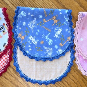 May include: Three baby bibs with scalloped edges. One bib features robots on a white background with a red gingham bottom. Another is blue with a dinosaur pattern. The third is pink with hearts.