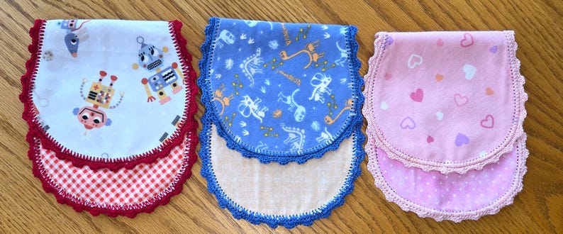 May include: Three baby bibs with scalloped edges. One bib features robots on a white background with a red gingham bottom. Another is blue with a dinosaur pattern. The third is pink with hearts.