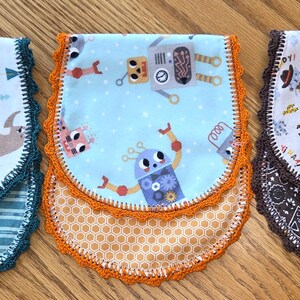 May include: Three baby bibs with different patterns and colors. One bib features sloths riding bicycles, another has robots, and the third showcases cowboys. Each bib has a scalloped crochet trim in coordinating colors.