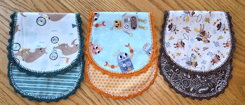 May include: Three baby bibs with different patterns and colors. One bib features sloths riding bicycles, another has robots, and the third showcases cowboys. Each bib has a scalloped crochet trim in coordinating colors.